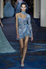 The image features a model walking down a runway, showcasing a glamorous dress. The dress is designed in a beautiful shade of blue and is adorned with intricate beadwork that catches the light. It has one long sleeve and a sculpted silhouette that highlights the model's figure. The background suggests an elegant fashion show setting, adding to the overall sophisticated atmosphere. The model completes the look with stylish footwear.