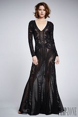 The image features a model wearing a striking black gown. The dress has a deep V-neckline and long sleeves, showcasing intricate detailing and patterns throughout. The material appears sheer in some areas, creating a layered effect. The design highlights the model's figure while exuding elegance and sophistication. The background is neutral, allowing the dress to stand out as the focal point.
