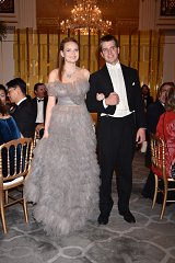 The image depicts a formal event taking place in an elegant setting, likely a gala or a high-profile gathering. In the foreground, a young woman wearing a stunning, light gray, layered tulle gown with off-the-shoulder sleeves stands beside a man in a classic black tuxedo with a white shirt and bow tie. They appear to be in a cheerful mood, possibly walking together through the venue. The background features lavish decorations, including a chandelier and a beautifully set dining area with gold chairs, indicating a luxurious atmosphere. Other guests can be seen seated at tables, enjoying the event.