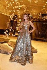 The image features a young woman wearing a stunning, shimmering gold gown. The dress has a sleek, fitted bodice with a deep neckline and a flared, flowing skirt that glimmers under the lights. She has a cheerful expression, standing confidently with her hands on her hips. The background suggests an elegant venue, with artistic decorations hanging from the ceiling, adding to the luxurious atmosphere. The overall scene conveys a sense of sophistication and celebration.