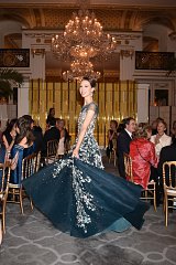 The image features a woman elegantly dressed in a stunning gown adorned with intricate floral embroidery. She is in a grand setting, characterized by opulent chandeliers and an elegant decor typical of a formal event. Guests are seated at tables in the background, some engaging in conversation while others are captivated by her graceful presence. The overall atmosphere is sophisticated and celebratory, highlighting the beauty of the gown and the venue.