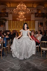 The image depicts a formal event taking place in an elegant venue, characterized by opulent decor, including a large chandelier and luxurious seating. In the foreground, a woman is wearing an ornate, silver-gray gown with intricate designs. She is smiling and gracefully holding out the skirt of her dress, showcasing its full and flowing design. The audience in the background appears to be engaged, with some applauding and others observing the woman with admiration. The overall atmosphere is one of celebration and sophistication.