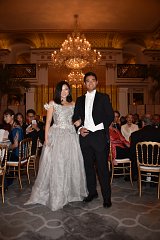The image depicts a sophisticated event, likely a formal gathering or gala. A couple is prominently featured in the foreground. The woman is wearing an elegant silver gown adorned with intricate details, while the man is dressed in a classic tuxedo with a white bow tie. The background shows a luxurious setting with ornate decor, chandeliers, and guests seated at tables, contributing to the refined atmosphere of the occasion. The lighting is warm and inviting, enhancing the opulence of the scene.