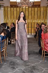 The image features a young woman walking confidently in an elegant setting. She is wearing a stunning gown that has a delicate pattern and flows to the floor. The venue appears to be a formal event, with golden decor and elegant seating. In the background, other guests are seated at tables, some of whom are looking in her direction. The overall atmosphere conveys a sense of sophistication and celebration.