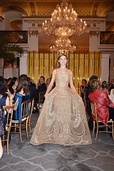 The image features a young woman wearing an elegant, flowing gown that is adorned with intricate beadwork and embroidery. The gown has a strapless design and a full skirt, giving it a glamorous and formal appearance. She is standing in a lavishly decorated indoor setting, likely during a formal event or celebration, with a large chandelier hanging above. In the background, several attendees are seated at tables, dressed in sophisticated attire, creating a festive atmosphere. The overall ambiance exudes elegance and luxury.