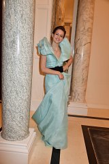 The image features a person in an elegant, light blue gown. The dress has a structured design with large, ruffled sleeves and a flowing skirt. The individual is standing gracefully next to a marble column, smiling. The setting appears to be a sophisticated indoor environment, possibly an event or gala. The overall vibe is one of glamour and style.