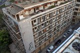 L'image montre un bâtiment abandonné, avec plusieurs étages visibles. La façade est en mauvais état, avec de la peinture écaillée et des balcons vides. On peut également apercevoir des traces de végétation sur le toit, suggérant un manque d'entretien. Des voitures garées le long de la route en contrebas ajoutent une touche de vie à cet environnement désolé. L'ensemble dégage une atmosphère de désuétude et de négligence.