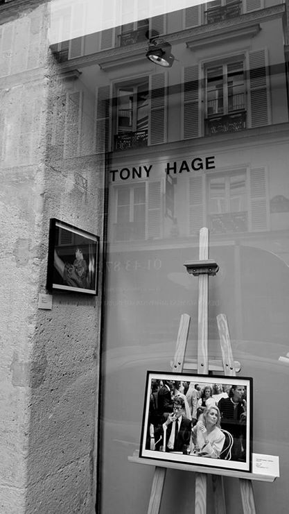 L'image montre une vitrine d'une galerie d'art avec des photographies exposées. On peut voir le nom "TONY HAGE" inscrit en haut. Deux photos sont accrochées : une est montée sur un chevalet, l'autre est fixée au mur. L'extérieur reflète certains détails d'un bâtiment voisin avec des fenêtres. L'atmosphère semble artistique et contemporaine, et l'image est en noir et blanc, ce qui ajoute une dimension nostalgique.