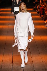 The image depicts a fashion model walking down a runway. She is wearing a sophisticated, all-white outfit that consists of a loose-fitting top with voluminous sleeves and a coordinated skirt. The outfit has a sleek and modern aesthetic, complemented by knee-high white boots. The model's hair is styled straight and smooth, and she has bold dark lipstick, adding a striking contrast to her look. The runway is well-lit, featuring warm orange lighting that enhances the overall atmosphere of the scene. The audience is visible in the background.
