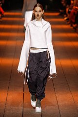 The image features a model walking down a runway during a fashion show. She is wearing a stylish, modern outfit that consists of a white cropped top with long, flared sleeves. The top has a slightly distressed look at the edges. She pairs it with loose-fitting, dark cargo pants that have drawstrings at the waist. Her footwear consists of chunky, gray shoes. The runway is lit with warm orange lighting, creating an atmospheric setting as the audience can be seen in the background. The overall look is contemporary and avant-garde.