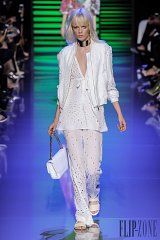 The image features a model walking down a runway wearing a stylish all-white outfit. The ensemble includes a lightweight, embroidered jacket with a delicate tie at the waist, paired with a matching top and flowing trousers. The model's long blonde hair and minimal accessories enhance the elegant look. The runway is set against a vibrant, colorful backdrop, creating a striking contrast with the outfit. The overall aesthetic is chic and modern, showcasing a sophisticated fashion statement.