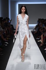 The image shows a model walking down a runway during a fashion show. She is wearing a white dress that features intricate floral detailing and a deep neckline. The dress has a thigh-high slit on one side, adding a modern touch to the elegant design. The model has long, sleek hair and is paired with white platform shoes. The background features an audience, suggesting a lively fashion event, with cameras and phones capturing the moment. The overall atmosphere conveys a sense of sophistication and style.
