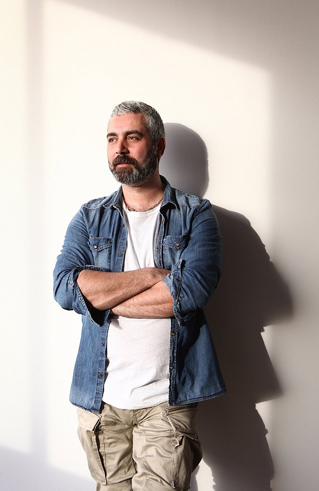 The image features a man with a beard and short gray hair, standing with his arms crossed in front of him. He wears a denim shirt over a white t-shirt and light-colored cargo pants. The background is a plain wall, illuminated by natural light, creating a soft shadow behind him. The overall mood appears relaxed and contemplative.