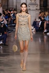 The image depicts a fashion model walking down a runway during a fashion show. She is wearing a shimmering, intricately detailed short dress that features elaborate embroidery and embellishments. The dress has a fitted bodice and a flared skirt, showcasing a blend of elegance and glamour. The model's accessories include a circular handbag and knee-high socks adorned with embellishments, adding to her stylish look. In the background, seated guests are observing the runway, creating a vibrant fashion show atmosphere.