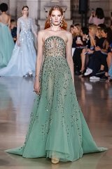 The image features a model wearing a stunning strapless gown in a soft green color. The dress is intricately embellished with delicate floral patterns and shimmering beads, creating an elegant and ethereal look. The model has long, flowing hair and is styled with a subtle makeup that complements the gown. In the background, other models are visible in similarly styled outfits, highlighting a fashion showcase or runway event. The setting appears to be a grand indoor space with a polished floor and soft lighting, adding to the overall glamorous atmosphere.