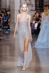 The image features a model walking down a runway wearing a glamorous evening gown. The dress is adorned with shimmering sequins and has a high slit, adding a daring touch to the elegant design. The gown appears to have a soft, light blue color that complements the model's style. She is accessorized with a delicate headpiece and wears matching embellishments on her legs. The background shows an audience admiring the fashion show, creating a lively atmosphere. The overall aesthetic conveys sophistication and high fashion.