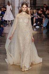 The image features a model walking down a runway wearing a stunning, sheer gown. The dress is adorned with intricate beadwork and delicate embellishments, giving it a shimmering effect. It has a form-fitting design that accentuates the model's figure and flows elegantly into the floor. The gown is complemented by a flowing, translucent cape that adds an ethereal quality. The model's hair is styled elegantly, possibly with a headpiece, creating a sophisticated and glamorous look. The background includes an audience, adding to the atmosphere of a fashion show.