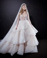 The image features a model wearing an elegant wedding gown. The dress has a layered design with ruffled tiers, creating a voluminous silhouette. It is adorned with intricate beading and features long sleeves with a delicate, sheer fabric. The bodice has subtle embellishments and the model is styled with a long veil that flows down from a decorative hairpiece. The background is dark, which contrasts beautifully with the light tones of the gown. The overall look is sophisticated and bridal.
