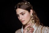 The image features a close-up portrait of a young woman with a thoughtful expression. She is adorned with elegant accessories, including a decorative headband and statement earrings that resemble wings. Her makeup is soft, enhancing her features, and her hair is styled in loose waves. The background is dark, which highlights the details of her outfit, embellished with pearls or similar decorative elements. The overall aesthetic is sophisticated and glamorous, suggesting a fashionable or high-end context.