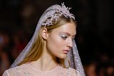 The image features a model showcasing an elegant and ethereal bridal look. She has long, flowing hair and is adorned with a delicate headpiece that resembles intricate, frosted branches or crystals. Her makeup is soft, with a subtle shimmer on her eyelids, enhancing her natural beauty. She is wearing a sheer, intricately designed gown with embellishments that likely complement the overall bridal theme, conveying a sophisticated and romantic aesthetic. The background suggests a runway setting, typical of a fashion show.