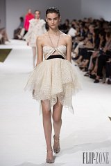The image features a runway scene during a fashion show. A model is walking down the catwalk, showcasing a stylish strapless dress made of delicate lace. The dress is short in the front, with a longer, flowing back. The model has long, sleek hair and is wearing sandals with embellishments. In the background, there are other models and an audience watching the show, with some people seated and taking photos. The overall atmosphere is vibrant and glamorous, typical of high fashion events.