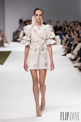 The image features a model walking down a runway, showcasing a stylish dress. The dress is light-colored with floral patterns and has puffed sleeves, creating a playful and feminine silhouette. The model's hairstyle is sleek, possibly pulled back, and she is wearing elegant accessories. The runway setting includes an audience in the background, highlighting the fashion event atmosphere. The overall aesthetic conveys a blend of sophistication and modern design.