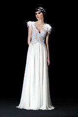 The image showcases a high-fashion gown characterized by its elegant design. The dress features a flowing white fabric that drapes gracefully to the floor, creating a soft and ethereal silhouette. The bodice is adorned with intricate beadwork and embellishments, adding a touch of glamour. Highlighting the shoulders are delicate feathers, which enhance the overall opulence of the gown. The model wears a sleek headpiece, contributing to the sophisticated aesthetic of the look. The background is dark, emphasizing the brightness and details of the dress.