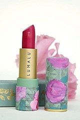 The image features a lipstick product. The lipstick itself is a vibrant pink, displayed in a gold tube with the brand name "LUMALU" printed on it. The outer packaging showcases a colorful design adorned with floral patterns, predominantly featuring roses and geometric shapes. In the background, there is a soft-focus flower that adds a touch of elegance to the scene. The overall composition emphasizes beauty and femininity.