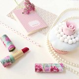 The image features a beautifully arranged flat lay. At the center, there's a white cupcake decorated with a pink rose made from icing. Surrounding the cupcake are several decorative elements, including a string of white pearls, a patterned notebook with a pink and white design, and a pink-striped straw. There are also two lipsticks with floral designs, one of which is opened, revealing a bold red color. The overall aesthetic is soft and feminine, enhanced by pastel colors and elegant details.