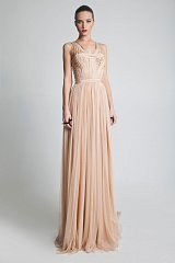 The image features a model wearing a long, elegant gown in a soft peach or nude color. The dress has a fitted bodice with intricate detailing and a flowing, sheer skirt that cascades to the floor. The model's hair is styled neatly, and she has a polished makeup look with bold lips. The background is neutral, allowing the dress to stand out as the focal point.