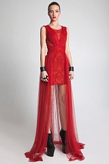 The image features a model wearing a striking red dress. The dress has a fitted design with intricate detailing, showcasing a blend of lace and sheer fabrics. It has a high neckline and is sleeveless, accentuating the model's arms. The skirt is short in the front and extends into a long, flowing sheer overlay at the back. The outfit is complemented by black, chunky platform heels and bold accessories, contributing to a dramatic and fashionable look. The background is plain and light, highlighting the vibrant red of the dress.