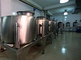 The image shows a row of large, shiny stainless steel tanks or containers situated in a clean, well-lit kitchen or food processing area. The tanks are elevated on sturdy metal stands, and some have valves or gauges attached. The background features tiled walls and overhead lights, suggesting a hygienic environment, likely used for food preparation or storage.