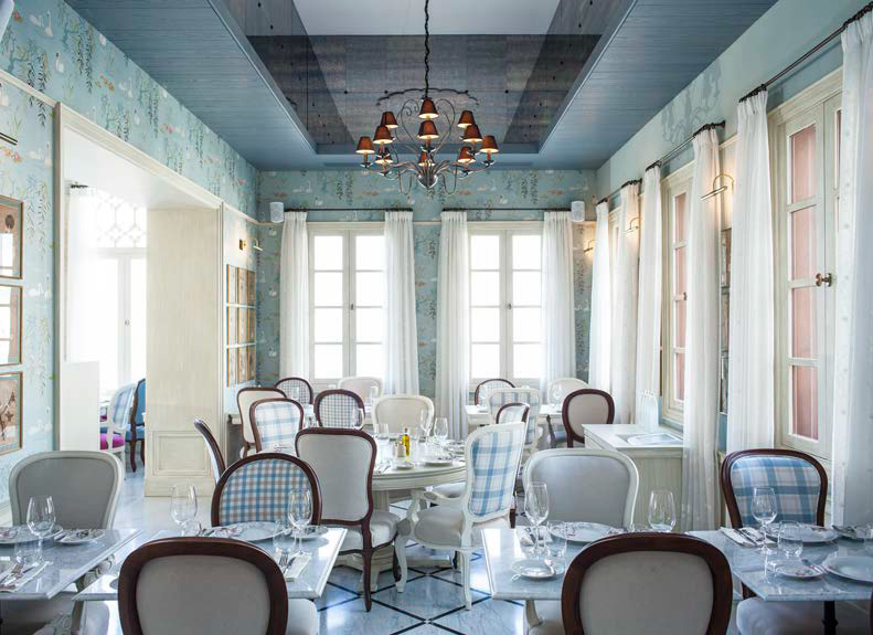 The image depicts a stylish and elegant restaurant interior. The walls are adorned with patterned wallpaper, and the space features large windows that allow natural light to flood in, enhancing the inviting atmosphere. The dining area consists of several round and square tables, each set with fine dining tableware, including glasses and utensils. The chairs are a mix of solid colors and patterned upholstery, contributing to a refined yet cozy feel. A unique chandelier hangs from the ceiling, adding a touch of sophistication to the overall decor. The light color palette, combined with tasteful furnishings, creates a calm and pleasant dining environment.