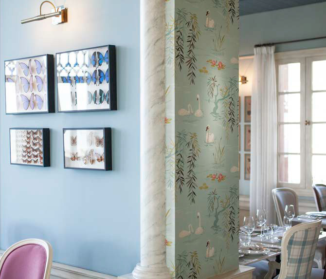 The image features a well-decorated interior space, likely a dining area or restaurant. One side shows a pale blue wall adorned with framed artworks depicting butterflies, arranged in a grid. Adjacent to this, there is a column painted in a light marble texture, which supports the ceiling. The column leads to another wall that is covered in a patterned wallpaper featuring swans and decorative foliage. The overall atmosphere appears elegant and inviting, with soft lighting and a blend of stylish furnishings in the background, suggesting a refined dining experience.
