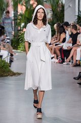 The image features a model walking down a runway during a fashion show. She is wearing a white, hooded dress that has a relaxed fit and a belted waist. The dress is knee-length and has short sleeves. The model is also wearing platform sandals with a chunky heel. The runway is set in a lush, green environment with an audience visible in the background, suggesting a vibrant fashion event. The overall look is casual yet stylish, combining comfort with a contemporary design.