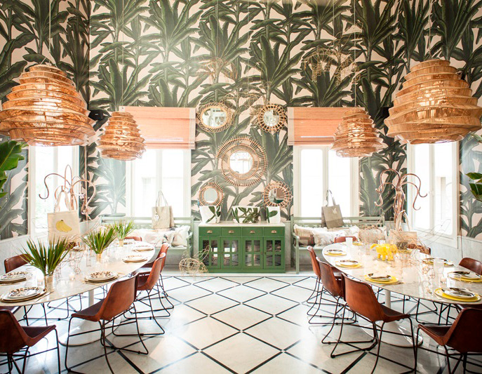 The image features a stylish dining area with a tropical theme. The walls are adorned with lush, green palm leaf wallpaper, creating a vibrant, leafy atmosphere. The room is brightly lit by natural light coming through large windows, which are adorned with woven shades. Central to the space is a circular dining table surrounded by modern, leather chairs. The table is elegantly set with plates and glassware, suggesting an inviting dining experience. Hanging above are unique, large pendant lights made of woven materials, adding a touch of warmth and texture. Decorative mirrors and circular wall art complement the overall design, while potted plants bring a bit of nature indoors. The flooring features a bold, geometric pattern, enhancing the contemporary feel of the space.