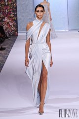 The image features a model walking down a runway, showcasing a glamorous light blue gown. The dress has a sleek, satin finish and is designed with a deep neckline and a high slit on one side, creating an elegant silhouette. One shoulder is adorned with a draped detail, adding an element of sophistication. The model has a polished hairstyle and is wearing high heels, complementing the overall refined look. The runway setting includes a floral backdrop, enhancing the fashion display.