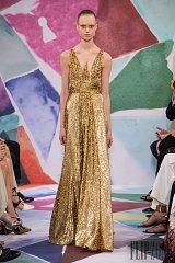 The image features a model presenting a glamorous gold dress. The dress is long and shimmery, characterized by a fitted bodice with a deep V-neckline and flowing skirt. The fabric appears textured, reflecting light beautifully. The background includes colorful geometric patterns, suggesting a vibrant setting, while seated audience members in the foreground hint at a fashion show environment.