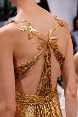 The image depicts a close-up view of the back of a dress. The dress is made of shimmering gold fabric, featuring intricate detailing and embellishments. The shoulder straps are designed with elegant, leaf-like shapes that are also gold in color. The flowing style of the fabric creates a graceful drape, enhancing the overall luxurious appearance of the gown. The setting appears to be a fashion show or fitting environment, with some blurred figures in the background.