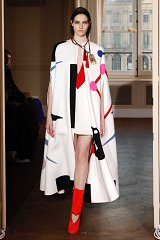 The image features a model walking down a runway wearing a striking outfit. She has on a white cape adorned with abstract patterns in various colors, including red, black, and pink. Underneath, she is dressed in a short, simple dress. The ensemble is complemented by bold red knee-high boots that have heart-shaped cutouts. The setting appears to be an elegant room with large windows, adding to the fashion show's sophisticated atmosphere.