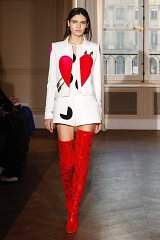 The image shows a model walking down a runway. She is wearing a bold and striking outfit that consists of a white tailored jacket featuring large red heart shapes and black accents. The ensemble also includes high-waisted shorts, which are complemented by eye-catching, knee-high red boots. The setting appears to be a fashion show, with natural light coming through large windows in the background. The overall look is vibrant and artistic, emphasizing a contemporary fashion aesthetic.