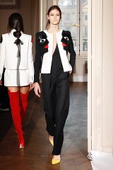 The image features a model walking down a stylish indoor runway. She is wearing a striking outfit that combines a black jumpsuit with a white jacket that has bold, artistic features, including red accents and embellishments. Her footwear consists of bright yellow heels, which add a pop of color to the ensemble. Another model can be seen in the background, wearing a similar jacket but with a different color scheme, highlighting a fashionable and chic atmosphere. The setting appears elegant, with large windows allowing natural light to fill the space.