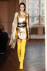 The image features a model walking down a runway wearing a striking outfit. The dress is made of a shiny, metallic gold fabric and has a fitted silhouette that reaches mid-thigh. The model accessorizes with a wide belt decorated with intricate designs. She also wears bold, knee-high yellow boots that add a vibrant pop of color to her look. The background shows an elegant interior, characterized by large windows and soft natural light, creating a sophisticated atmosphere for the presentation.