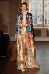 The image features a model walking down a runway, showcasing an elegant outfit. She is wearing a long, flowing dress with a shiny, gold material that creates a luxurious appearance. The dress has a deep neckline and is complemented by a colorful, richly embroidered jacket adorned with intricate patterns. The setting appears to be a stylish indoor location, possibly during a fashion show, with large windows in the background allowing natural light to illuminate the scene. The model's hairstyle is sleek and modern, contributing to the overall sophisticated look.