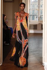 The image features a model walking on a runway, showcasing a striking dress. The dress is long and flowing, made from a sheer black fabric adorned with vibrant, colorful floral and geometric patterns. It has a deep neckline and features ruffled sleeves, adding a dramatic touch to the ensemble. The background suggests a well-lit, elegant setting, possibly from a fashion show, with an audience visible in the background. The model's posture is confident, embodying the essence of high-fashion presentation.