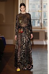 The image features a model walking down a runway wearing a long, flowing dress with a sheer overlay. The dress has an intricate pattern that combines dark tones with colorful accents, giving it a dynamic and eye-catching appearance. The model's hair is styled neatly, and she carries a small, decorative handbag. The setting appears to be an elegant indoor space, with large windows that allow natural light to filter in, highlighting the details of the dress.