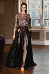 The image features a model on a runway wearing an elegant outfit. The top is transparent with intricate embroidery, showcasing vivid colors and patterns. The skirt is long, flowing, and made of sheer fabric, featuring a high slit that reveals the model's leg. The overall look is dramatic and stylish, complemented by striking orange shoes. The setting appears to be a fashion show, with a sophisticated indoor backdrop.
