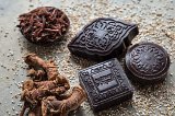 The image features a variety of chocolate squares with intricate designs, laid out on a textured surface. Surrounding the chocolates are small piles of spices, including strands of dried herbs and root pieces, likely contributing to the overall aesthetic and flavor profile. The earthy tones of the ingredients complement the rich color of the chocolate, creating an inviting and organic appearance.