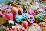 The image features a colorful assortment of handmade soaps arranged in a vibrant display. The soaps are various shapes and sizes, including heart shapes and circular ones, with swirls of colors like pink, blue, and yellow. Many soaps are individually wrapped in clear plastic with decorative red ribbons. In the background, there are more soaps, suggesting a rich variety of styles and colors. Overall, the image conveys a lively and artistic presentation of bath products.