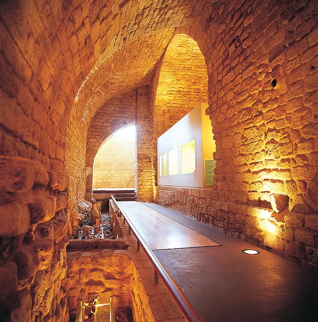 The image depicts a historic interior space featuring impressive stone arches and walls. The stonework is textured and warm in color, illuminated by soft lighting that highlights the architectural details. A pathway or walkway runs through the area, leading towards displays or exhibits, with a glimpse of a lower section suggesting a multi-level design. Overall, the atmosphere conveys a sense of history and craftsmanship.